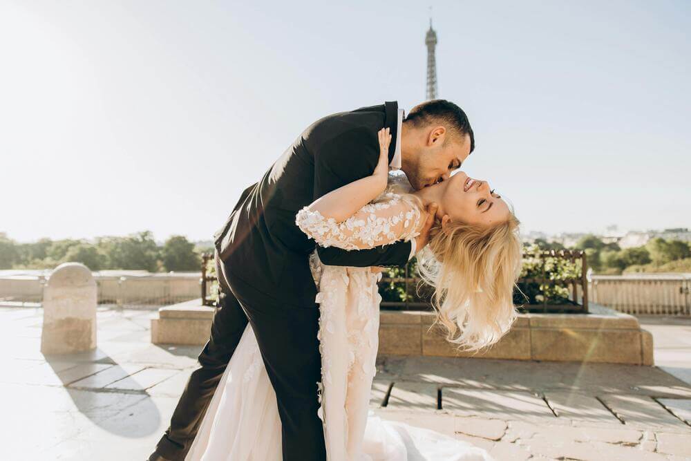 2024 Trending wedding styles and the shoes to match. Groom dipping his bride to kiss her on her neck in front of the Eiffel Tower on their wedding day.