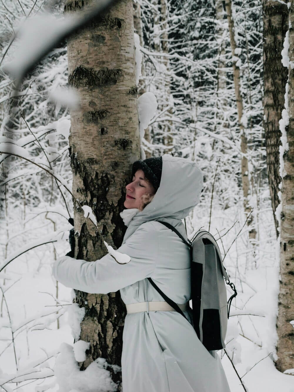 Woman in the snow hugging a tree and wearing a white rain coat and Rains Backpack. Our guide to Rains Backpacks: Your ultimate outdoor companion