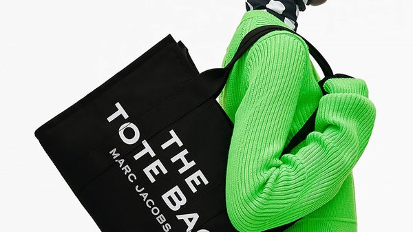 Our Top 10 Back-to-Office Bags for Women. Woman holding the Marc Jacobs The Black Canvas Tote Bag (large) wearing a black and white polka dot head scarf, neon green jumper and jeans.