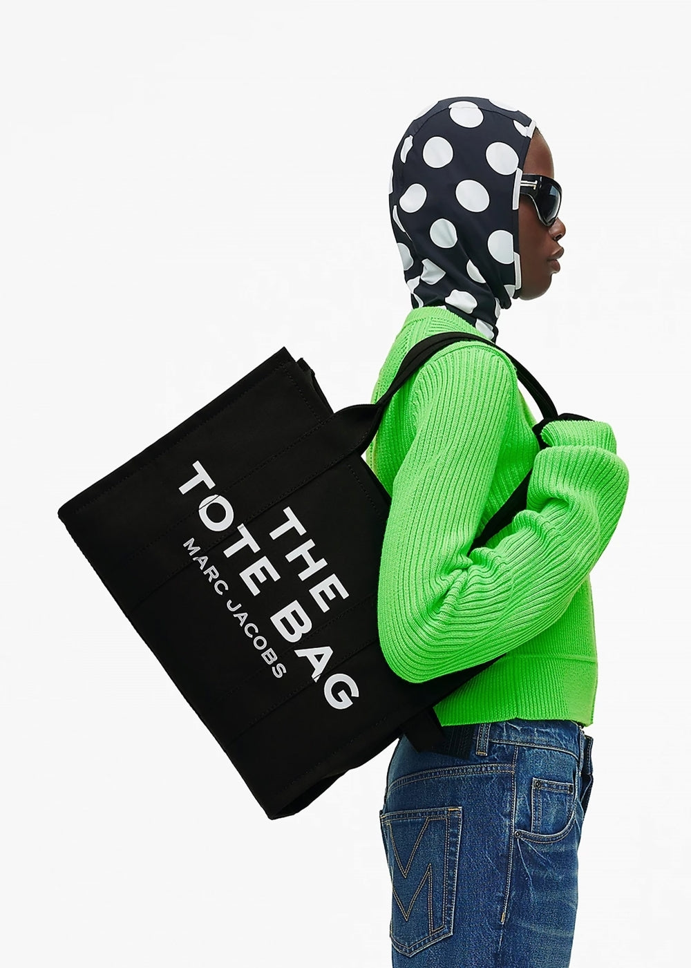 Our Top 10 Back-to-Office Bags for Women. Woman holding the Marc Jacobs The Black Canvas Tote Bag (large) wearing a black and white polka dot head scarf, neon green jumper and jeans.